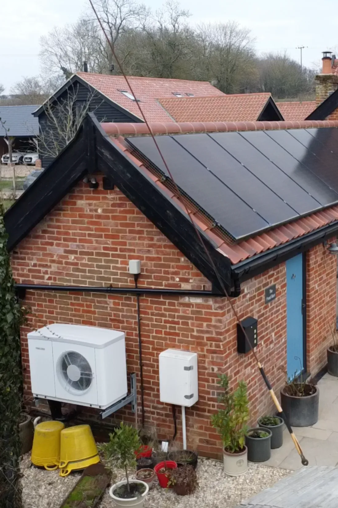 A red-brick outbuilding with solar panels on its roof and a white heat pump on the wall beside a blue door