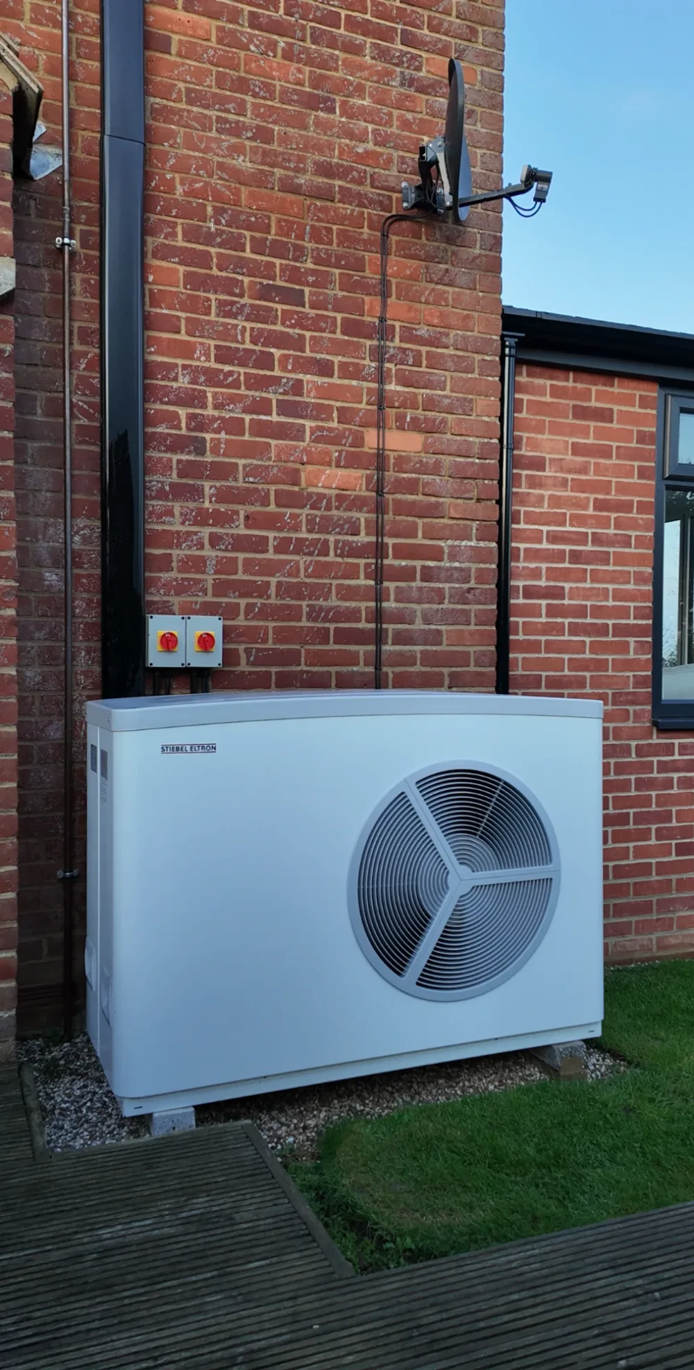 A Stiebel Eltron heat pump on a red-brick wall beside two isolators, with a satellite dish above