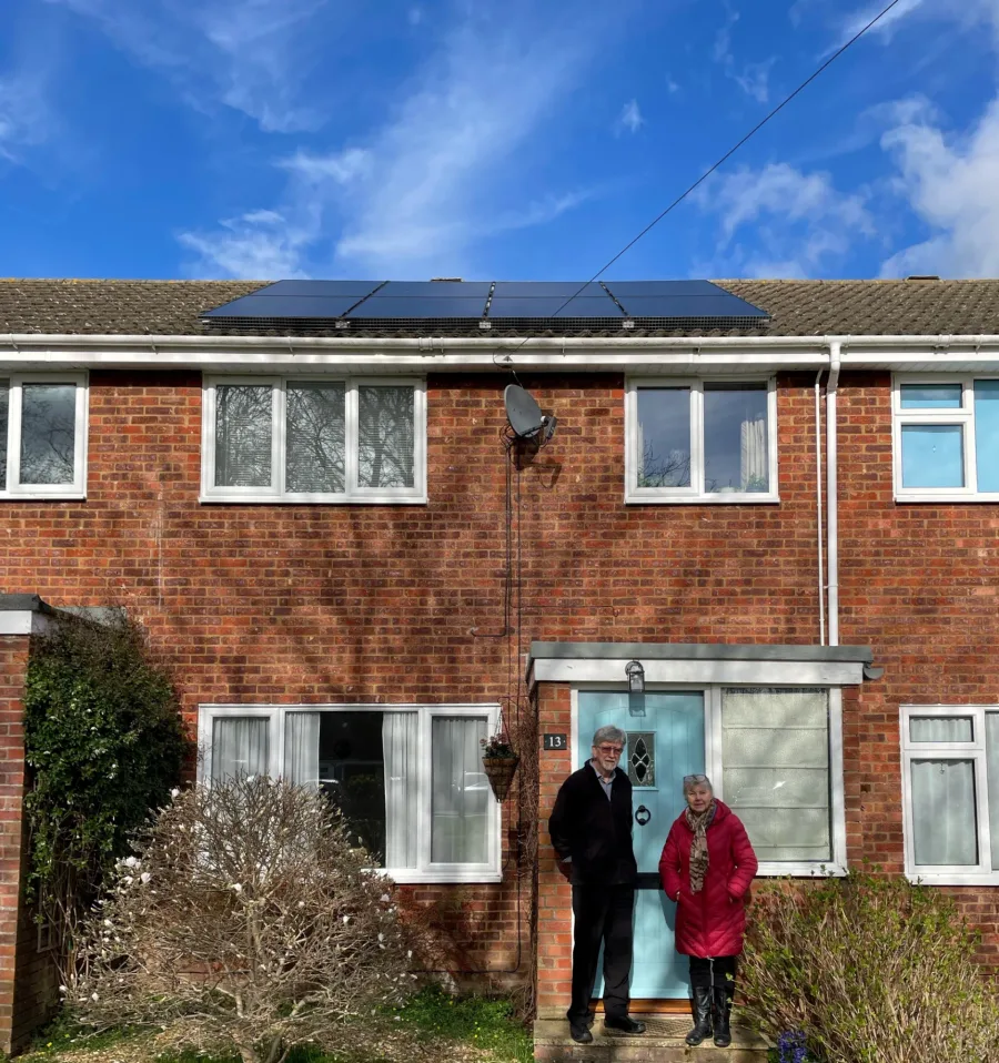 A couple standing at the pale blue front door of their red-brick terrace, with solar panels on the roof