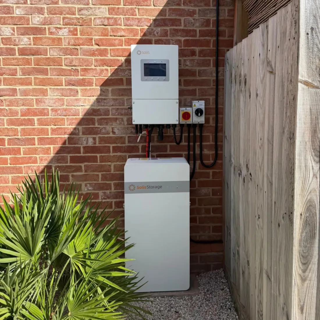 A Solis inverter above a Solis Storage battery on a red-brick wall beside a fence